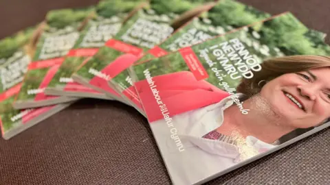 A set of manifestos laid out of a surface with Eluned Morgan's face on the booklets. The manifestos says "Pennod Newydd i Gymru".