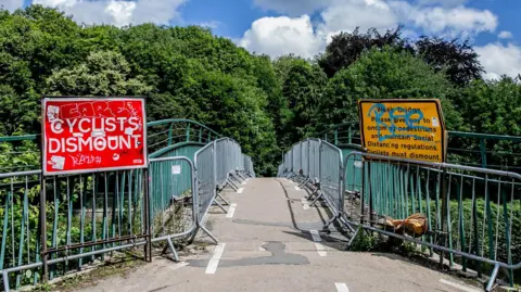Baths Bridge which is a curved concrete path with green metal railings on both sides. There are silver temporary barriers lining the bridge in front of the railings and two large signs at the beginning of the bridge. The one on the left is red and reads 'Cyclists dismount', while the one on the right is yellow and reads: 'Weak bridge please give way to oncoming pedestrians and maintain social distancing regulations. Cyclists must dismount'. They have both been defaced with graffiti. There are white broken up lines signalling cycle lanes on either side of the bridge. Over the bridge there are lots of green trees.