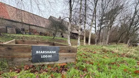 Brian Farmer/BBC A grey slate sign, reading "BEARSHANK LODGE" stands in front of a line of silver birch trees a long stone building with a red-tiled roof.