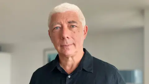 David Rogers is wearing a black buttoned shirt, with the top button undone, and has white, short hair.