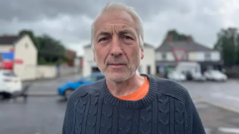 A man with white hair, navy jumper looking at the camera.