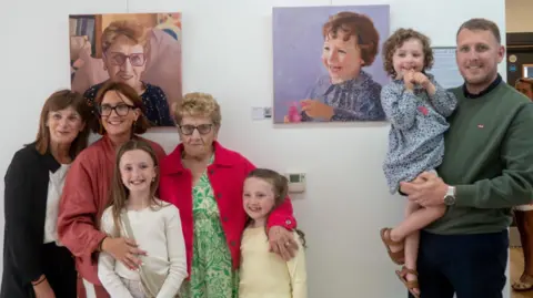 Photographs by Sara Two children and three women, including Victoria Smyth and Mary Brown stand in front of Mary's portrait on the wall. Beside them to the right is Hatty's portrait. Standing beside that portrait is a man holding Hatty in his arms. 