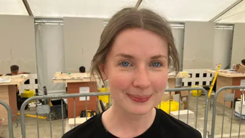 Rebecca Gavigan is wearing a long sleeved black top, has blue eyes and tied up brown hair. She is standing in front of wood work stations.