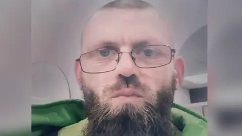 North Wales Police A man with black-framed wire glasses; he is close shaven and has a long brown beard. He is wearing a green jacket.