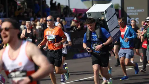 Getty Images Runner Jordan Adams participates in the marathon while carrying a full-sized 25kg white fridge strapped to his back to raise funds for frontotemporal dementia research.