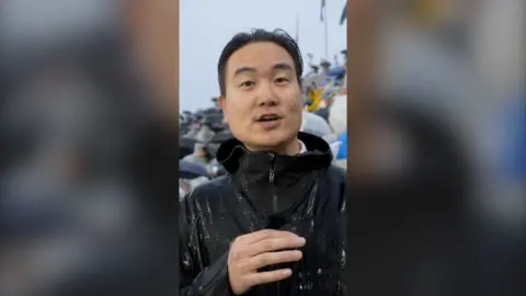 A BBC reporter in a raincoat surrounded by South Korean fans with umbrellas at the match