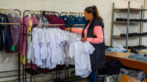 KDM Photography Pia Honey is looking through a rack of children's school shirts in a warehouse. Other clothing can be seen behind and there are also racks of shoes