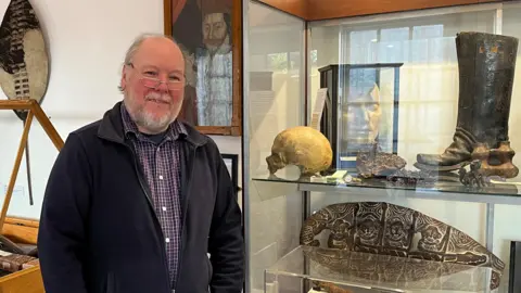 BBC Robert Bell, the museum's curator, is wearing a black fleece and checked shirt and standing next to a glass cabinet. Inside the cabinet are items including a skull, a large boot and a metal engraved item in the shape of an oval.
