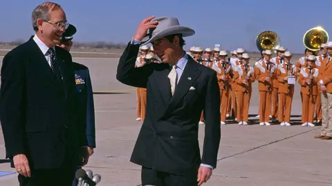 AP Charles dons a cowboy hat given to him by the mayor of Austin, Texas, during a visit to the city in 1986. A large brass band plays on the airport tarmac behind him