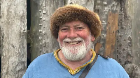 A man with a white beard, weather a blue top and a fur hat. 