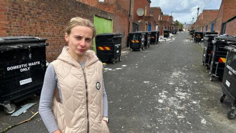 Blond haired woman in cream coloured parka stands in rubbish-laden road
