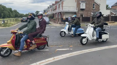 BBC/Louise Fewster Three moped scooters going round a corner. The first scooter is red and gold and being ridden by a man wearing a green jacket and a black helmet with a teenage boy wearing a dark purple suit and a silver helmet sitting on the back. Behind are two scooters - one white and one blue and white - being ridden by men wearing dark green jackets and blue jeans and helmets.
