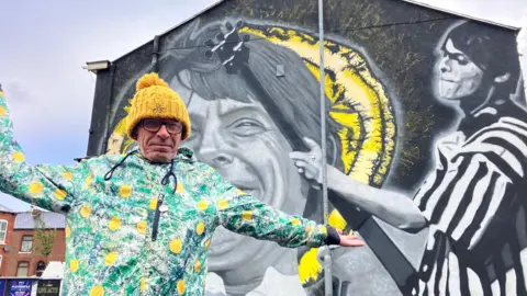 A man in his 60s wearing a yellow knitted bobble hat, glasses and a colourful anorak with lemons on the front stands in front of a large mural on the side of a building. 