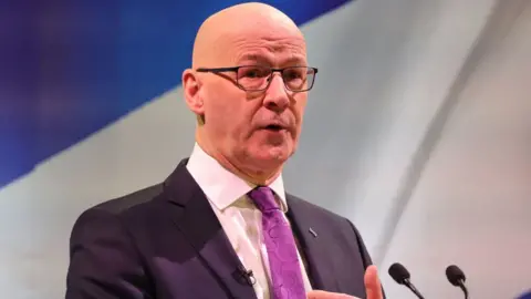 PA Media John Swinney, who is bald and wearing glasses, stands at a podium in front of a saltire. He is wearing a dark suit, white shirt and purple tie.