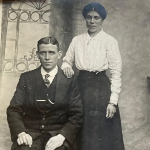 Supplied A black and white photograph of man sat in a three-piece suit, with a side parting in his hair, and a woman in a white shirt and long black dress stood behind his shoulder, with her hand resting on him