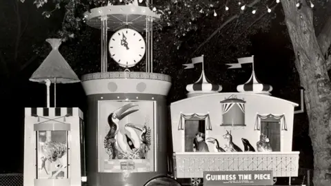 Maurice Mead Photography A black and white image of the guinness clock in the 1950s at the light festival. The vertical timepiece has a clock on top with painted toucans below. Next to it is a model caravan with flags on top and a model zoo keeper leading animals thorugh a door.