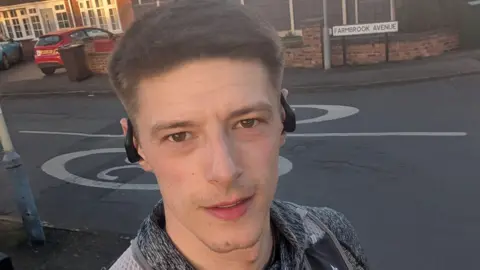 A selfie of a man with short brown hair, wearing black ear phones, a rucksack and a grey top. He is standing at the side of a residential street. A street sign is behind him as is a house with a red car in the driveway. 