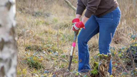 Holly Wilkinson A woman, wearing blue jeans and a brown jumper, with grey and red gardening gloves on, leaning into a garden fork, in a nature reserve with grass, plants and hedges all around her. You can only see her legs and top of her body. 