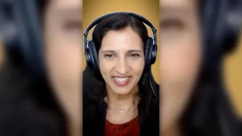 A woman with long dark hair, wearing radio headphones and a red top, smiles into the camera.