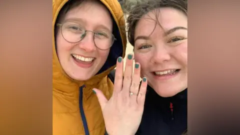 Ella Cook, pictured left, smiles at the camera. She is wearing glasses and a yellow puffer jacket with the hood up. Her fiancé, Kai Poa-Tams, is on her right. Kai smiles at the camera and is showing her engagement ring on her hand.