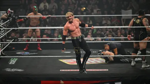 Getty Images Logan Paul is hanging of the top rope of a wrestling ring looking towards the crowd. There are four wrestlers and a referee behind him in the ring. Three of them are wearing masks. There are rows of people in the crowd behind the ring. 