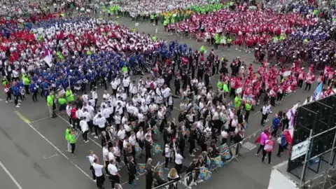 An aerial shot of hundreds of athletes in their colourful tracksuits standing in their team groups on a stretch of grey tarmac