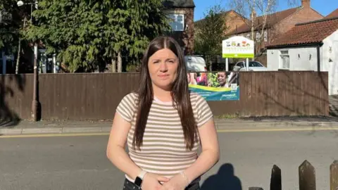 A woman frowns as she stands on the side of a narrow road outside a nursery. She has long brown hair and wears a white and brown striped T-shirt, dark jeans and a smartwatch with white strap. The nursery is behind a brown wooden fence with a sign reading "Town & Country Nursery". Cars can be seen in a small car park next to a white-washed building.
