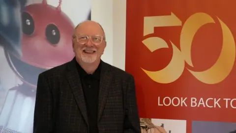 Shaun Whitmore/BBC John Gordon-Saker standing against a sign saying 50 on the right, while behind him is a poster showing a Martian robot used in the Cadbury For Mash get Smash ad. John is wearing glasses, a brown checked jacket over a black shirt and has a short white goatee beard.