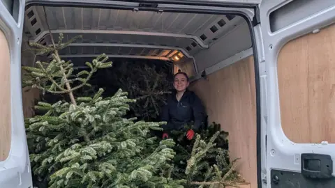 FORGET ME NOT CHILDREN'S HOSPICE A woman in a black coat in the back of a van with a green fir tree wedged against her.