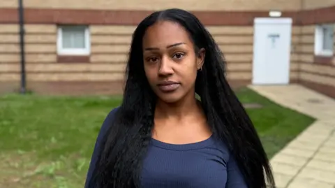 The head and shoulders of a woman with long black hair and a dark blue top standing outside a bricked building.