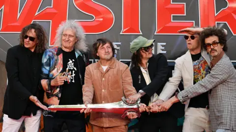Five men standing next to each other holding a silver sword prop, in front of a movie premier sign with bold red writing