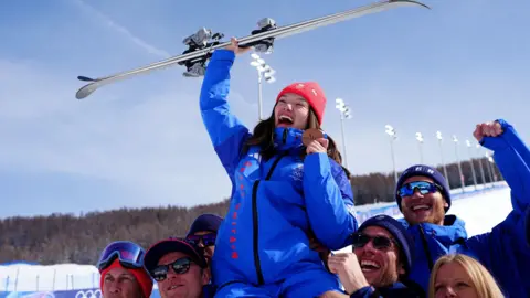 PA Media A woman in a blue Great Britain ski outfit and red woolly hat sits aloft the shoulders of six team-mates at the bottom of a ski slope. She is holding up a bronze medal in one hand and skis in the other. The slope is floodlit, although the picture is in blue skies and sunshine.
