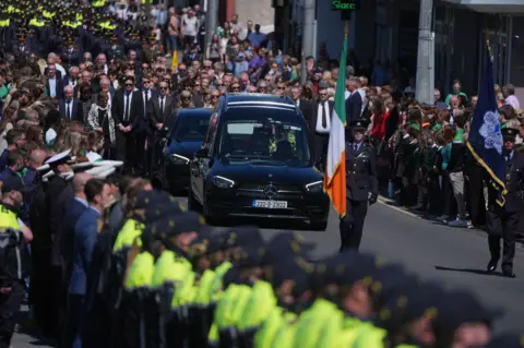 PA Media A large crowd line either side of the street as a black hearse drives down it, followed by a group walking. A man holding an Irish tricolor leads the procession.