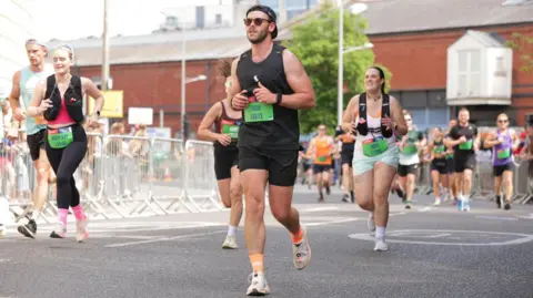 A man wearing black running clothes is running down a road. There are other runners alongside him.