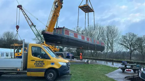 A yellow truck is in the foreground of the photo, in the middle of the image a red and black narrowboat is being lifted by a large yellow crane