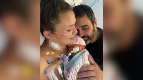 Kimberley Newark holds her baby in her arms, the baby is wrapped in a blanket and woollen hat and is connected to various medical tubes. The baby's father, Yann Trupiano is holding his face close to his daughters head with his eyes closed.