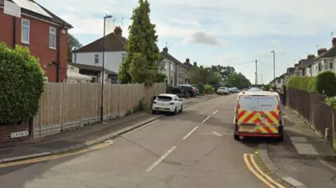 The entrance to a residential street, with houses either side and a number of vehicles parked partially on the pavement