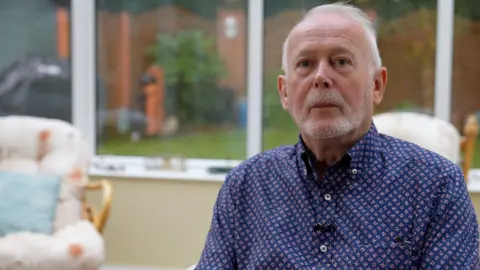A man in a pink and navy patterned shirt sits in a conservatory, a garden and arm chair seen behind him. He has white hair and a beard.