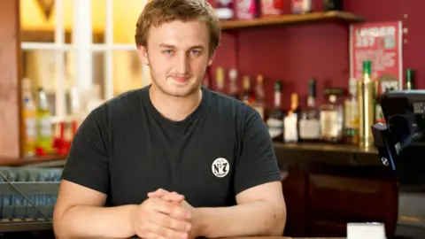 BBC Alex Barry, a young man with blond hair, stands behind an old fashioned bar in a pub.