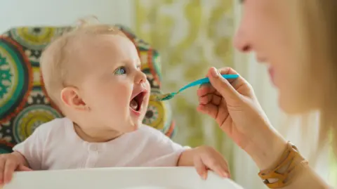 A stock image of a baby with blue eyes being spoon fed by their mother.