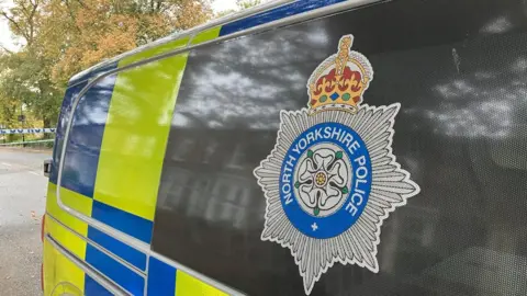 BBC The side of a North Yorkshire Police van with the force's logo on the it. Behind the van is an area of road with police tap across it and a tree.