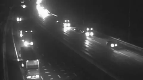 A black and white night time still from a highways camera. It shows the M25 near junction 29 showing lorries and cars with their lights on.