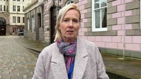  Catherine Curtis standing in front of a building with pink bricks. She is wearing a coat with a multi-coloured scarf. She has straight blonde hair.
