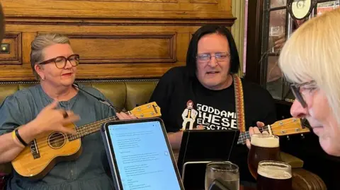 A woman with grey hair tied back is wearing a denim dress and is strumming her ukulele. She is wearing glasses and is reading music notes from an iPad. She is sitting next to a man with short black hair, also playing a ukulele. 