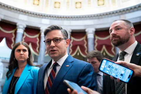 US House Speaker Mike Johnson surrounded by people and a phone held out, recording him. 