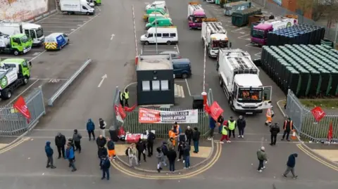 PA Media An aerial view of people standing outside the metal gates of a bin lorry depot. There are signs hung up on the fences and some people are in orange fluorescent clothing. On the right hand side there are two white bin lorries waiting to leave the depot, but people are blocking their way