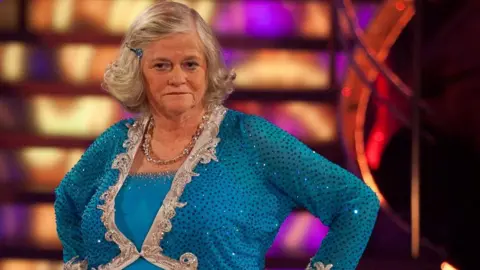 Ann Widdecombe stands on a brightly lit television studio floor wearing a blue sequinned costume with silver trim and a matching necklace. Colourful stage lights and illuminated set panels are visible in the background.