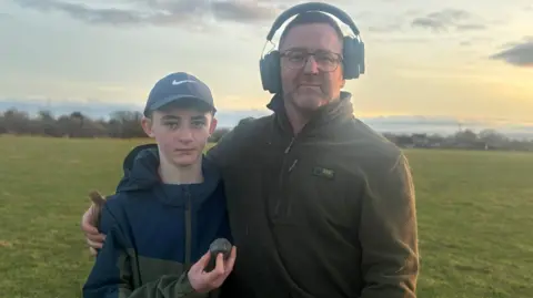 Dave Cunliffe is wearing headphones with his arm around his son Lucas who is holding a sword pommel in his hand. They are standing in a field with warm orange light on them. Lucas is wearing a Nike cap.