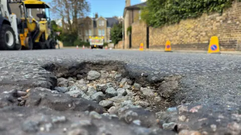 A pothole in the middle of a residential road. Rubble has been exposed due to the surface layer breaking off. The road has industrial machinery parked nearby and is lined with yellow cones.