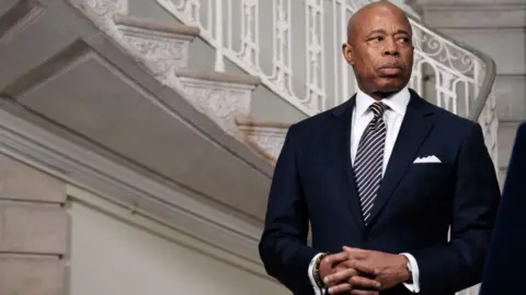 Eric Adams - a bald man - looks to the side, off camera. He is wearing a suit and there is a staircase behind him